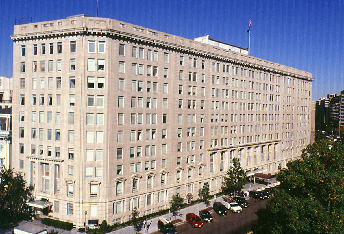 A long eleven-story limestone office building with rows of windows and an American flag on the roof, seen from above a street with parked cars under a clear blue sky