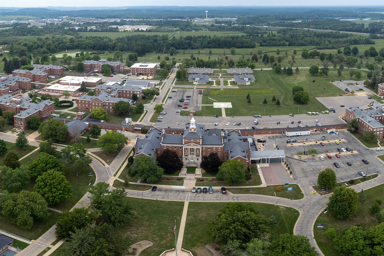 Aerial view of a campus of red brick buildings with a white cupola on the central building, parking lots, lawns and trees, with farmland and woods stretching to the horizon