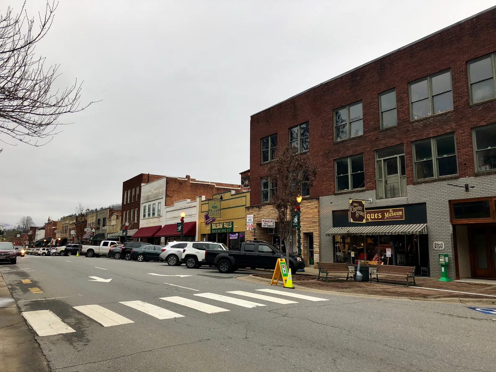 A small-town main street of two- and three-story brick buildings with shop awnings, a country store sign, benches on the sidewalk and cars parked along the curb under a gray sky