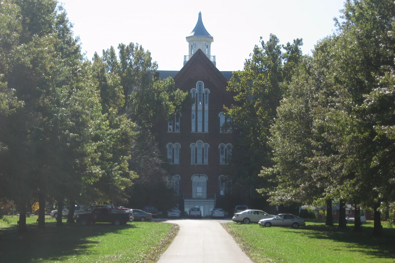A tall red brick building with arched windows and a white cupola stands at the end of a paved drive lined with trees and lawns, with cars parked in front of it