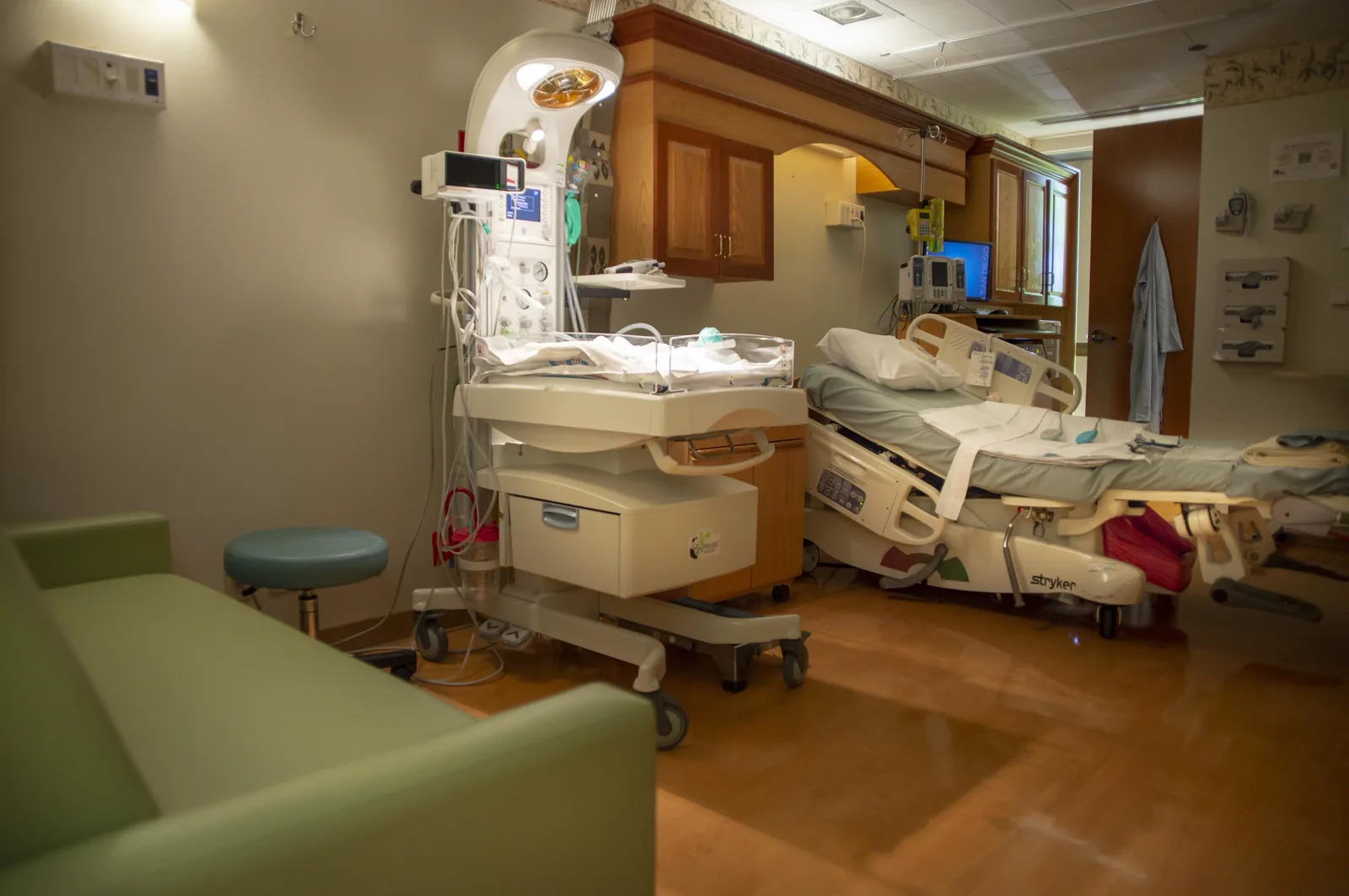 An empty hospital delivery room with an infant warmer and bassinet on wheels, a patient bed with monitors behind it, wooden cabinets and a green sofa in the foreground