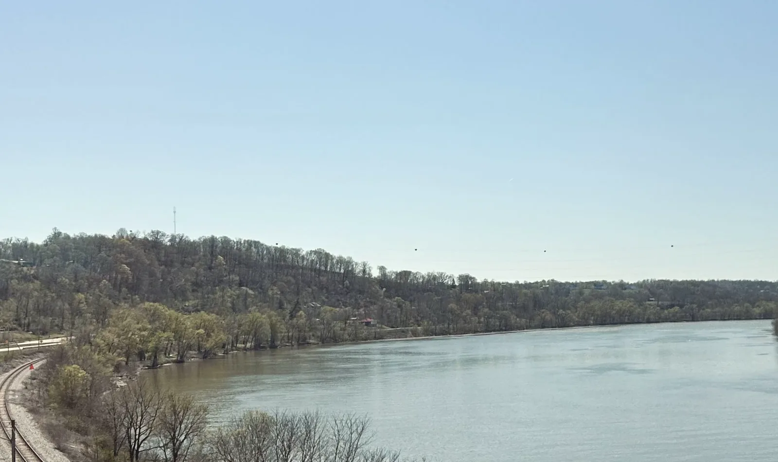A wide brown river curves between wooded hills under a clear blue sky, with a railroad track running along the near bank