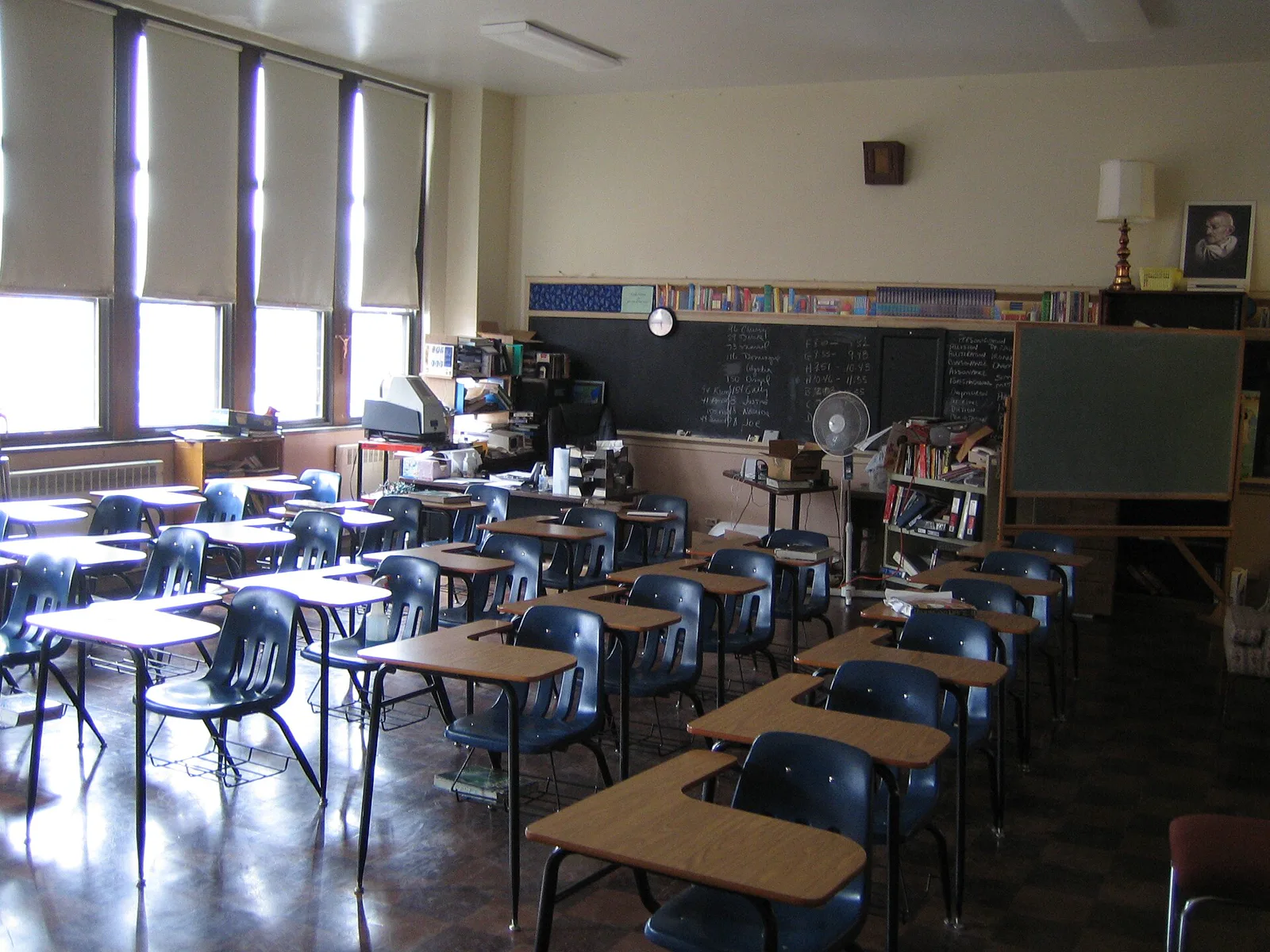 An empty classroom with rows of blue chairs and wooden desks, a chalkboard with writing on the far wall, shelves of books and tall windows with drawn shades